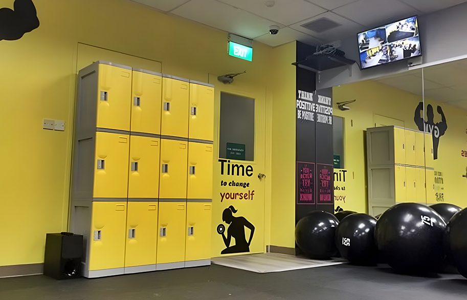Avios | Why ABS Lockers Are Gaining Popularity in Singapore’s Commercial Spaces A neatly arranged three-tier yellow locker system with gray frames in the corner of a fitness center (gym). Designed to securely store gym members' belongings, it showcases the durability and functionality of abs locker singapore products.